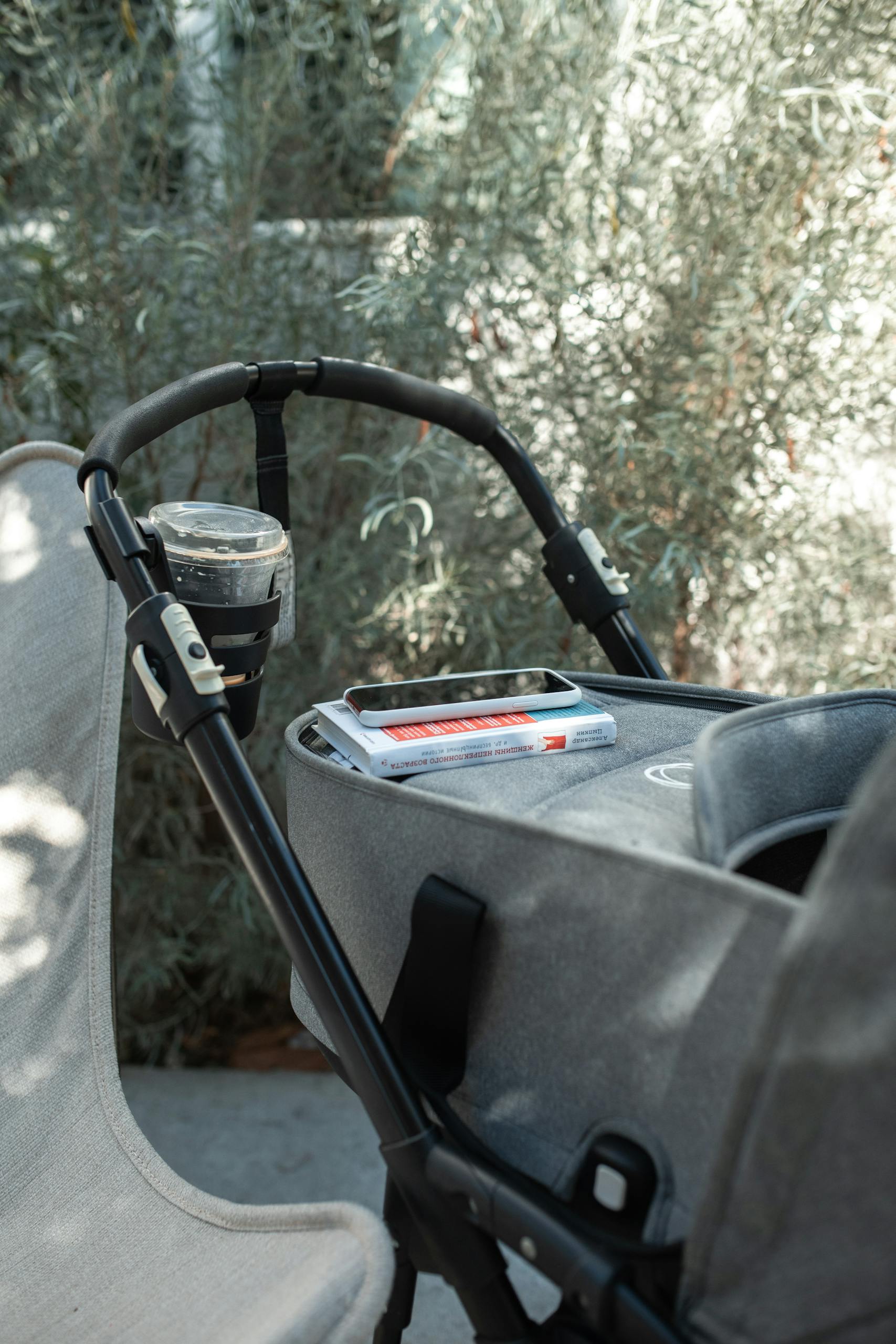 A baby stroller with a book and cup holder, set outdoors amid lush greenery.