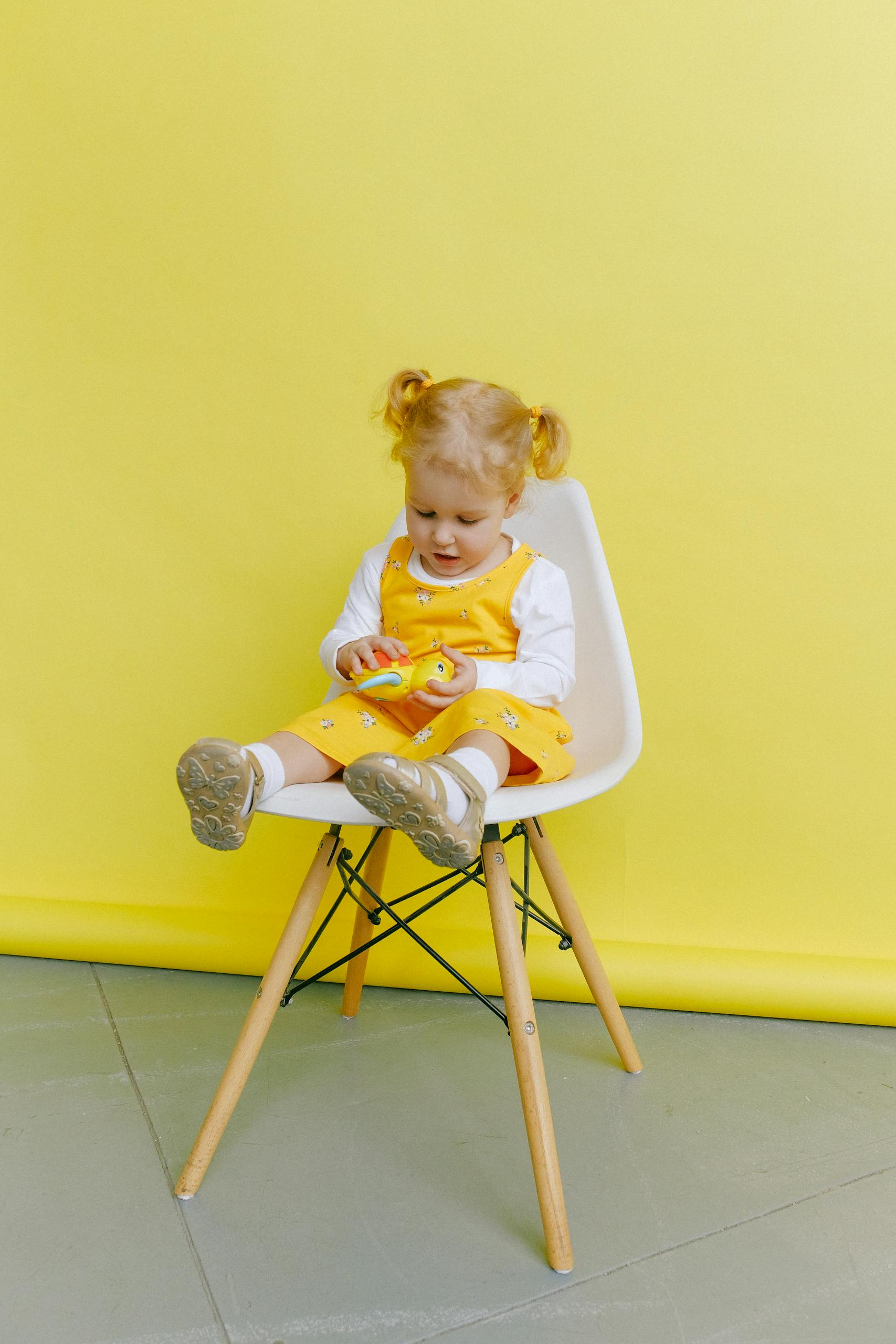 Adorable toddler girl in a yellow dress sitting on a chair against a bright yellow background.